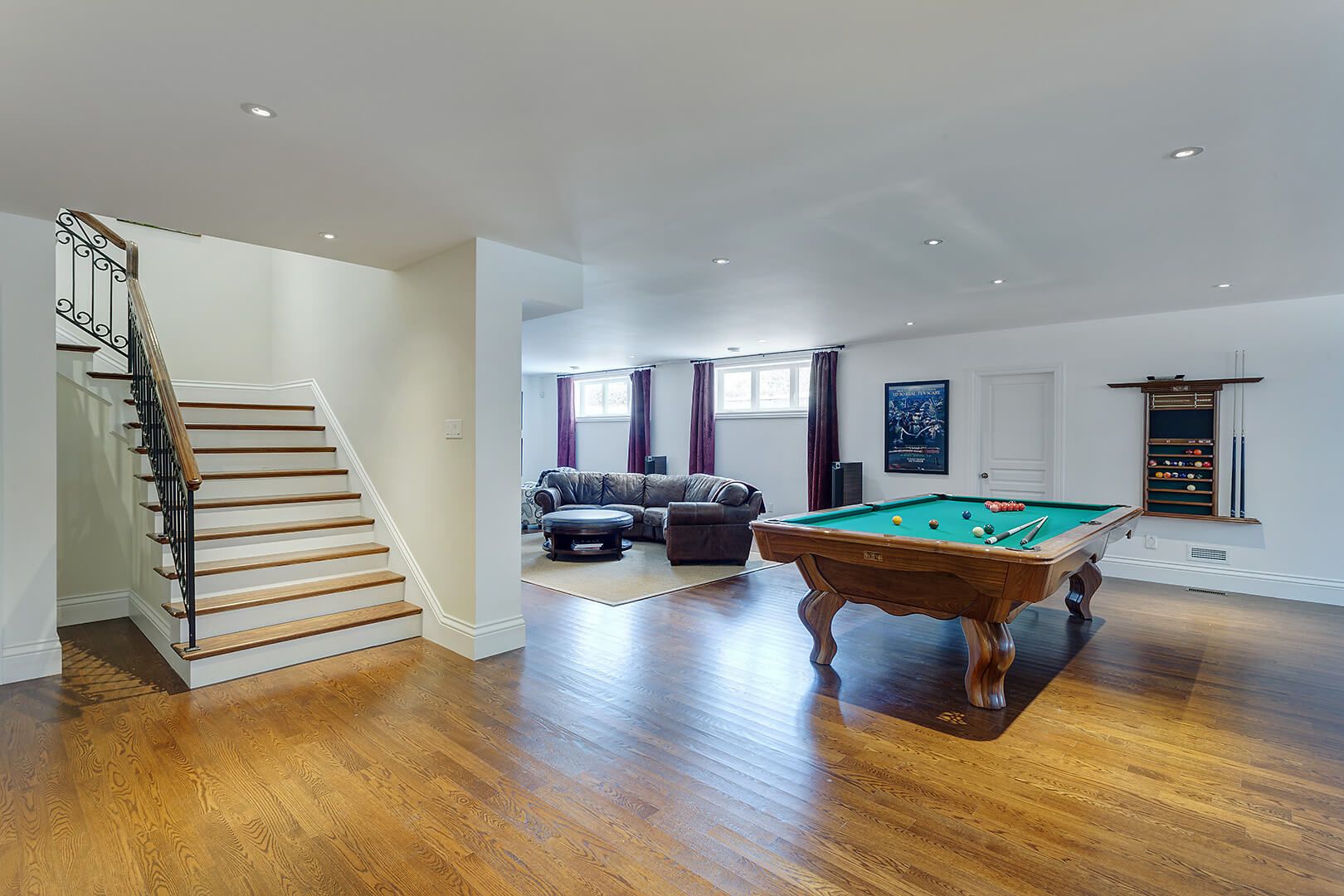 Finished basement featuring a billiards table, lounge area with leather sectional sofa, and wooden staircase.